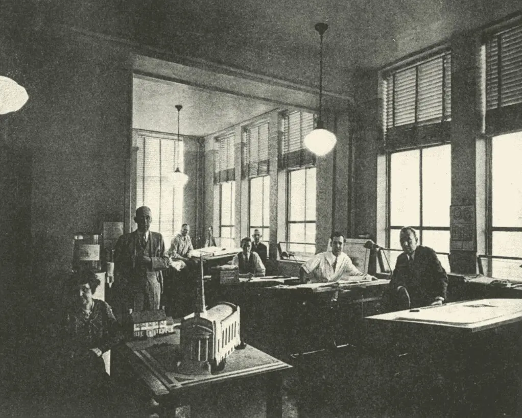 A group of seven people work at drafting tables in a well-lit room with large windows and overhead lights. Architectural models and blueprints are visible throughout the space.