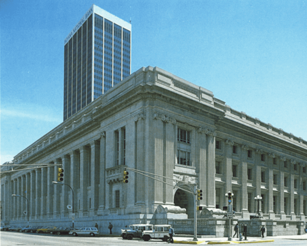 A historic, large concrete building with large columns stands on a street corner with modern skyscrapers in the background and a few cars and pedestrians at the intersection.